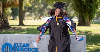 Woman wearing graduation cap and gown, jumping up in excitement in front of Allan Hancock College sign.