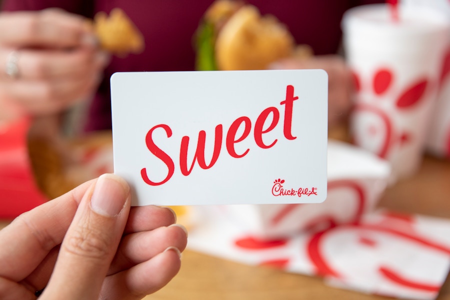 A person's hand holding a Chick-fil-A gift card in front of someone else eating a Chick-fil-A meal.