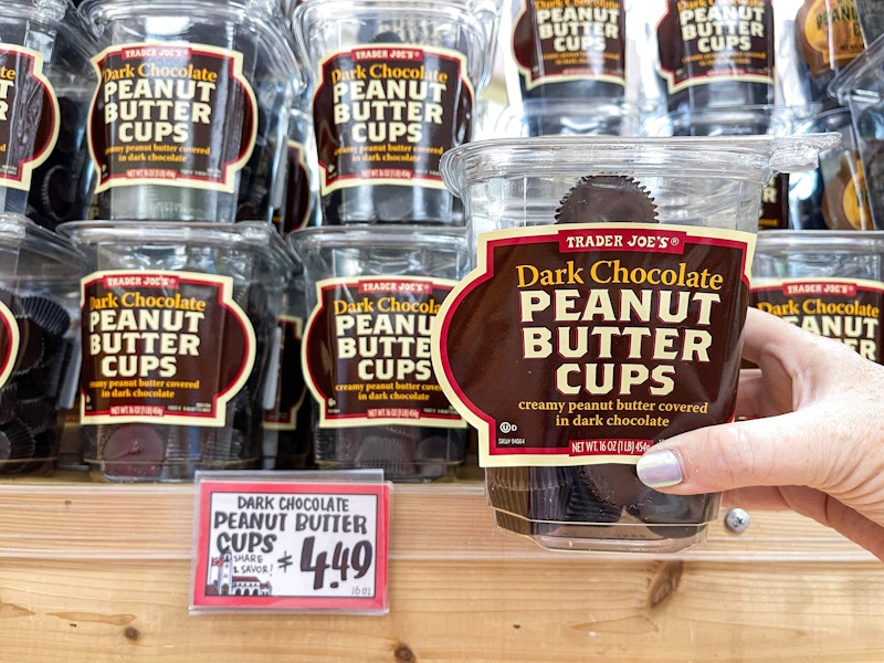 A person's hand holding a cup of Trader Joe's Dark Chocolate Peanut Butter Cups in front of a display of them on a shelf at Trader Joe's.