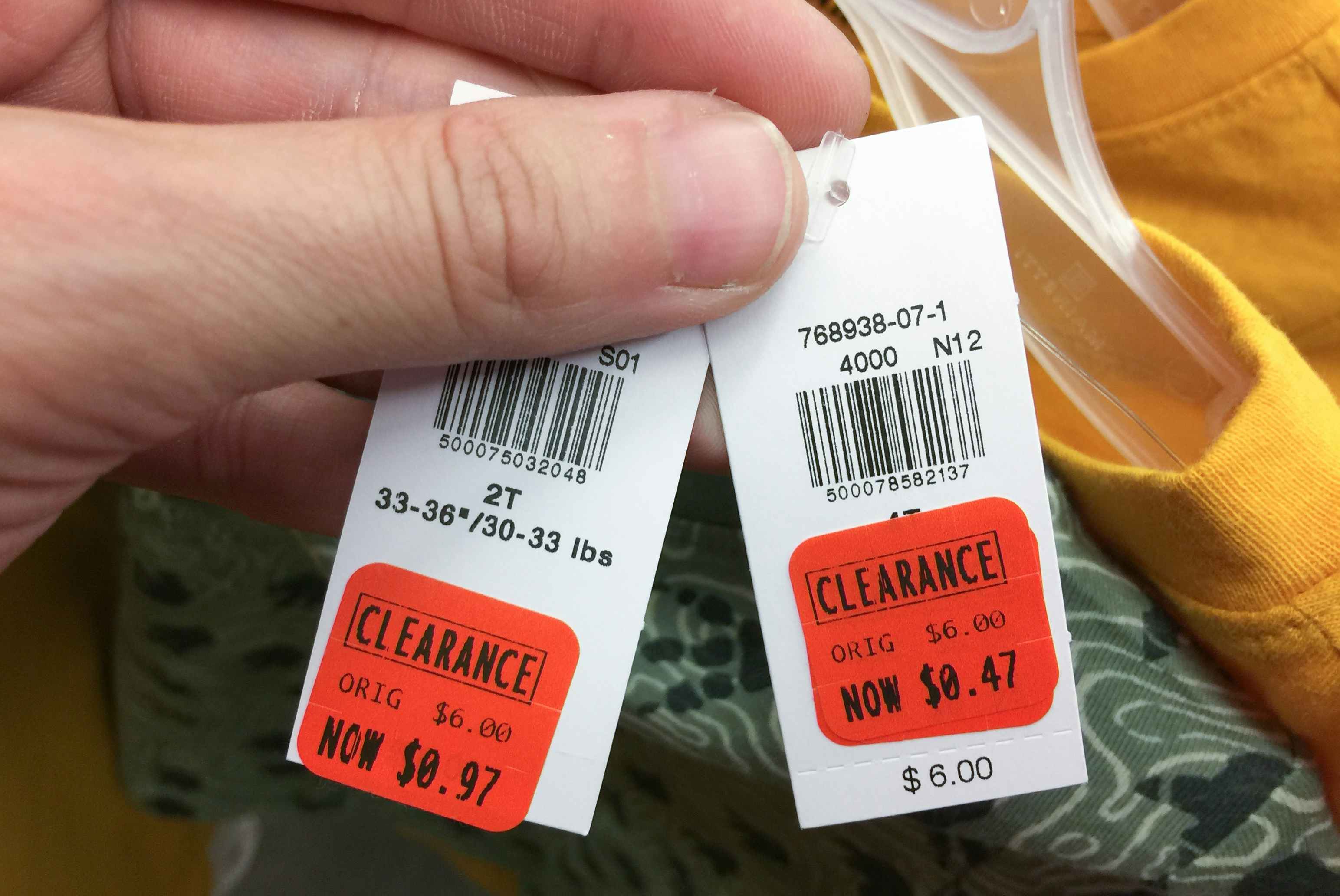 A person's hand holding up two clearance-stickered price tags, showing their marked-down prices, one being $0.97 and the other being $0.4...