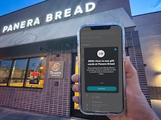 a person holding a phone up in front of a panera bread sign