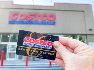 A person holding a Costco Card while standing in front of a Costco warehouse.