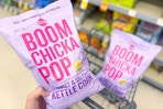 person holding a bag of boomchickapop popcorn over a cart with another bag of popcorn in it