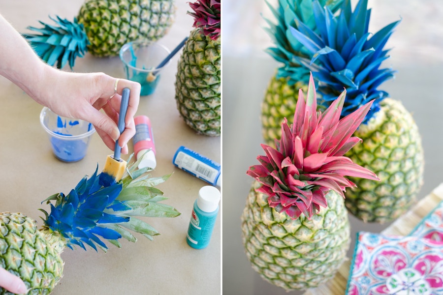 A person using acrylic paint to paint pineapple crowns next to some pineapples that have been painted pink and blue.