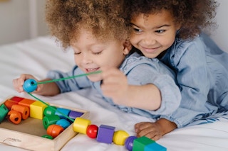 Two toddlers play with the Melissa & Doug Primary Lacing Beads on a bed.