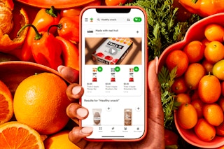 Hand holding phone with Instacart app opened in front of a background that has fresh fruits and vegetables