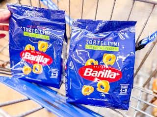 hand holding a bag of barilla tortellini pasta next to another bag in walmart cart