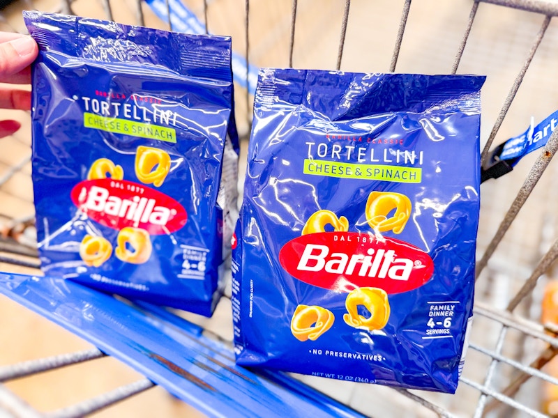 hand holding a bag of barilla tortellini pasta next to another bag in walmart cart