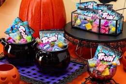 A Halloween snack table in orange, black and purple with pumpkins, spiders, and cauldrons full of Nerds, Trolli and Now and Later candy.