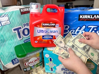 Someone holding some cash next to a stack of household staples including paper towels, toilet paper, laundry detergent, trash bags, and ...