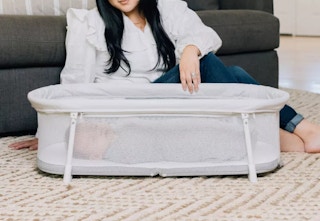 woman sitting next to a portable baby bassinet laying on the floor