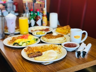 A served table at Denny’s with several items including a plate with eggs, sausage, and hash browns, a plate of an omelette and hash ...