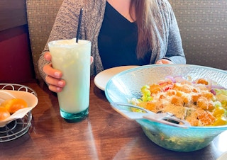 a woman holding a glass of Italian soda near a large bowl of salad 