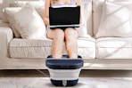 Lifestyle image of a woman using a foot bath while sitting on the couch using her laptop