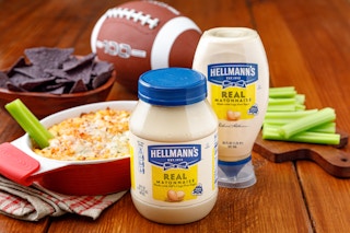 Hellmans mayo on a table next to a buffalo dip, celery, chips, and a football 