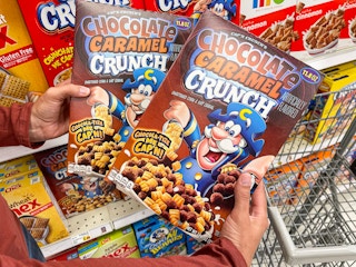 A person's hands holding two boxes of Cap'n Crunch's Chocolate Caramel Crunch in front of a shelf in the cereal aisle of a grocery store.