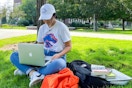 A woman sitting with books and a backpack and laptop on the lawn of a university campus.
