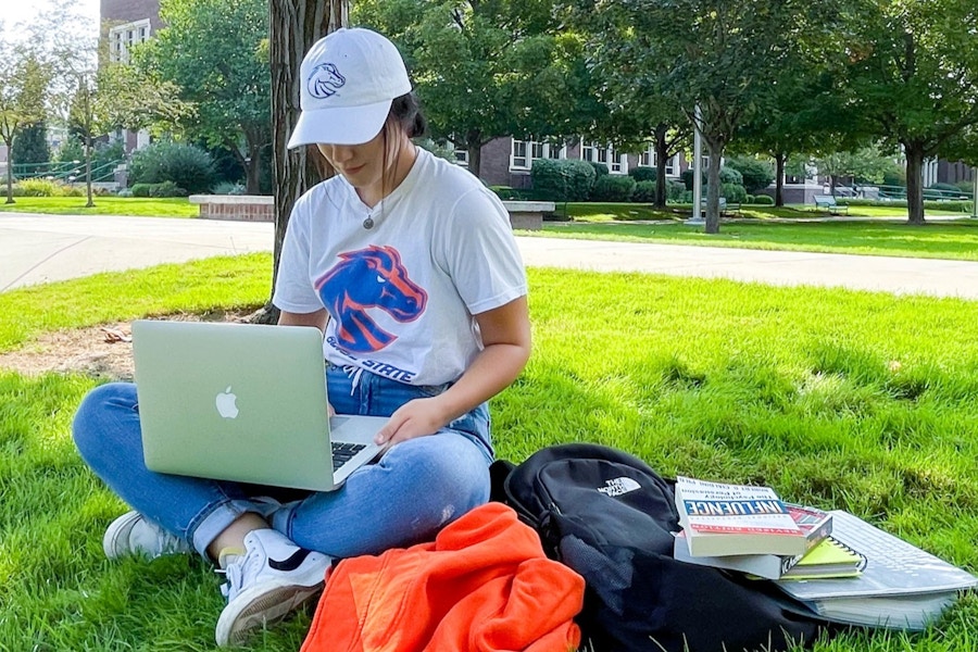 A woman sitting with books and a backpack and laptop on the lawn of a university campus.