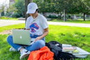 A woman sitting with books and a backpack and laptop on the lawn of a university campus.