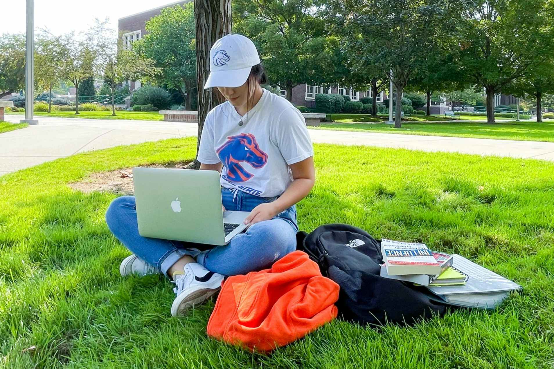 A woman sitting with books and a backpack and laptop on the lawn of a university campus.