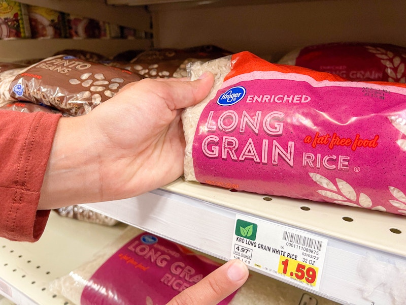 A person's hands, one holding up one end of a bag of Kroger enriched long grain rice sitting on a shelf, and the other hand pointing at t...