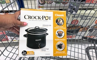 A hand holding a Crock-Pot in a shopping cart.