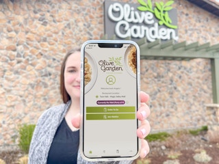 a woman holding out a cellphone in from of an olive garden with the waitlist app on phone