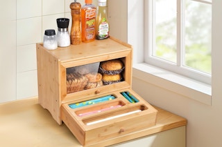 A bread box sits on a counter in the corner of a kitchen. Its bottom drawer is open to show bag storage.