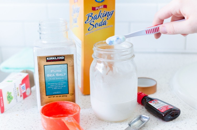 tooth brush dipped in mason jar filled with homemade tooth paste. Baking soda, vanilla, and sea salt on the counter beside.