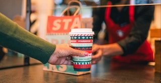A person grabbing a Starbucks cup at the counter with a barista in the background wearing a mask.