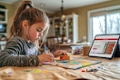 A young girl painting a craft at a kitchen table with an open ipad with Michaels online crafts on screen. 