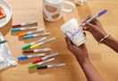 colorful Sharpie markers on a table under someone drawing on a cup 