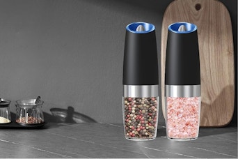 On a grey counter by a  grey wall with a wood cutting board against it are two grinders, one with pink salt, one with rainbow peppercorns.