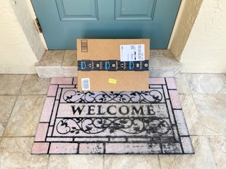 An Amazon box sitting on a welcome mat and leaning against a front door.