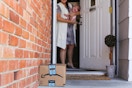 A mom holding a baby, opening the front door to an Amazon delivery box on the porch.