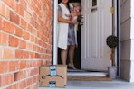 A mom holding a baby, opening the front door to an Amazon delivery box on the porch.