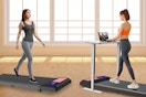 Two women walk on treadmill walking pads. One has a standing desk with a laptop on it. Both wear grey stretch pants.