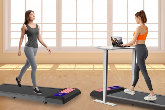Two women walk on treadmill walking pads. One has a standing desk with a laptop on it. Both wear grey stretch pants.