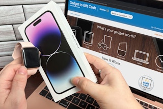 Someone holding an iPhone 14 and Apple Watch in front of a laptop displaying the Walmart electronics trade in webpage