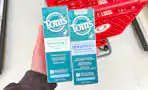 hand holding 2 tom's of maine toothpaste in front a parked target cart