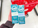 hand holding 2 tom's of maine toothpaste in front a parked target cart