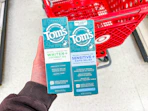 hand holding 2 tom's of maine toothpaste in front a parked target cart