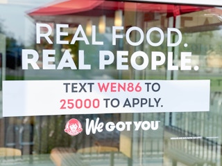 A window cling in a Wendy's window that reads, "Real Food, Real People. Text WEN86 to 25000 to apply. We got you.