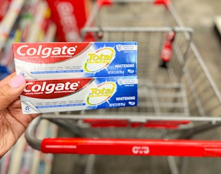 hand holding two tubes of Colgate toothpaste in front of shopping cart
