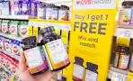 person holding 2 bottles of vitamins by a buy one, get one free sign
