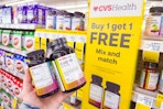 person holding 2 bottles of vitamins by a buy one, get one free sign
