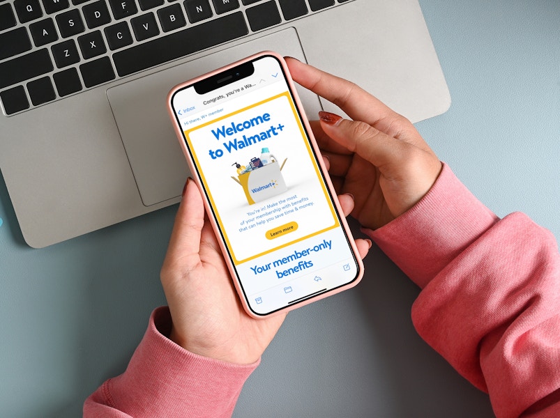 a woman's hands resting on a desk on top of a laptop, holding a phone and looking at a walmart plus welcome email