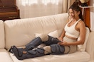 A young woman sits on a beige sofa with both legs wrapped in the leg compression massager. She holds the controller.
