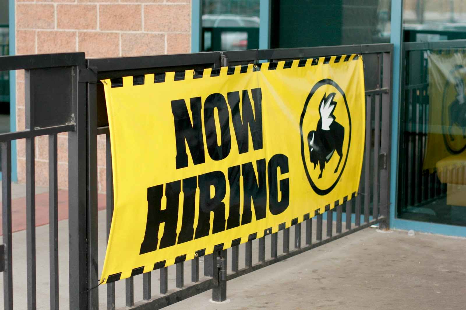 A bright yellow sign affixed to the fence outside of Buffalo Wild Wings which reads 