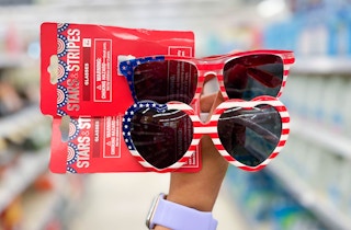 hand holding a pair of Stars & Stripes eyeglasses & heart eyeglasses in aisle